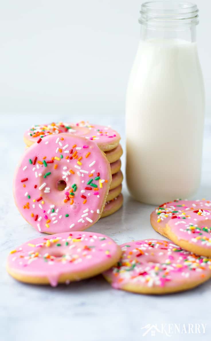 a stack of cookies shaded and decorated like pink donuts with sprinkles on top with a container of milk