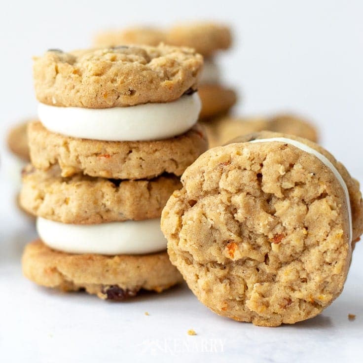 An oatmeal raisin carrot cookies is standing vertically on end so you can see the chunks of minced shredded carrot. Next to it on the left there are two carrot cake cookies stacked on top of one another. Each cookie is actually cream cheese frosting layered between two oatmeal raisin cookies like a cookie sandwich.