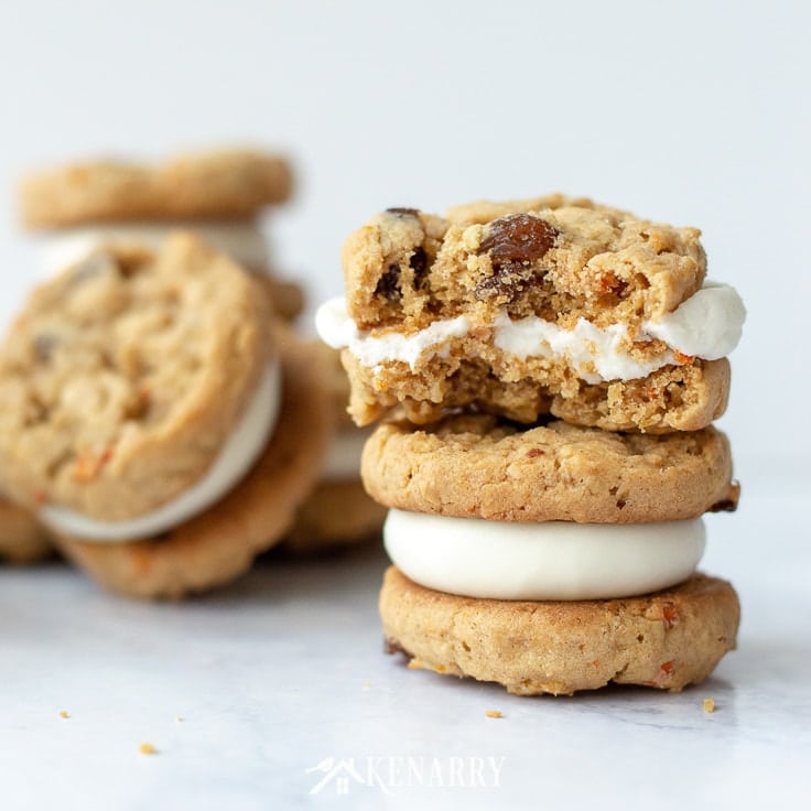 Two oatmeal raisin carrot cookies stacked on top of one another. These carrot cake cookies have cream cheese frosting in the center. The top cookie has a large bite out of the side of it.