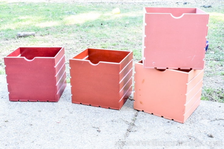 4 wood crates sitting outside. A red crate, orange create and 2 pink crates. 