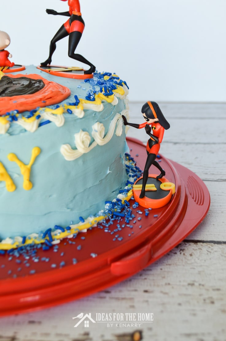 A toy action figure of Violet from The Incredibles appears to creating a force field at the side of a round double layer birthday cake