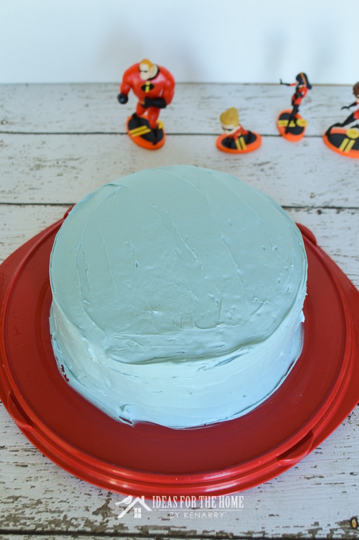 Round double layer cake covered with blue frosting on a cake carrier. Incredibles figurines are in the background.