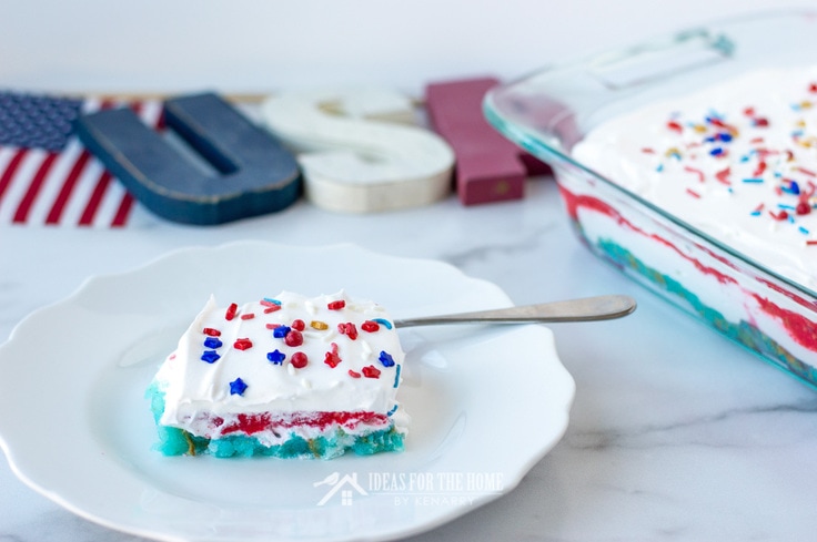 A slice of cake topped with whipped topping and sprinkles. The cake has layers of red white and blue for Independence Day.