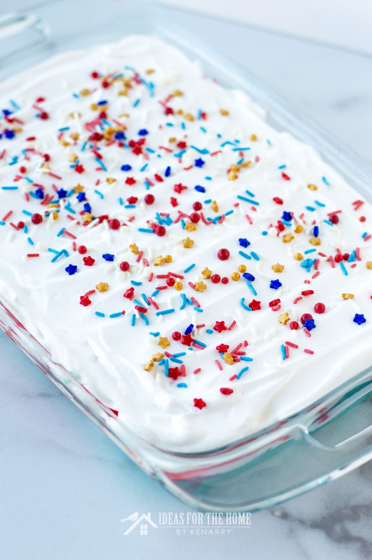 Overhead shot of a 9 x 13 inch glass pan filled with a layered jello cake covered in whipped topping and sprinkles for a 4th of July barbecue